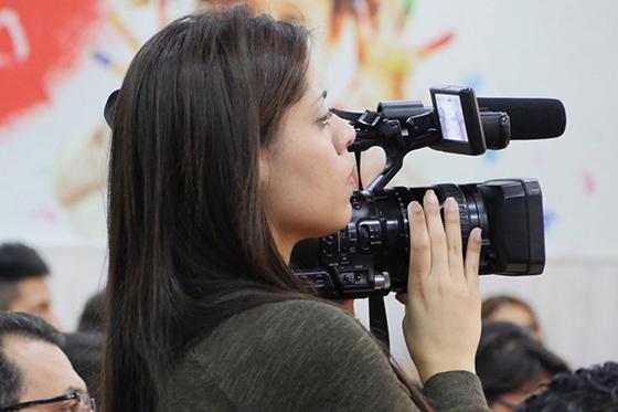 Photo of a woman looking into viewfinder of a video camera resting on her shoulder. 