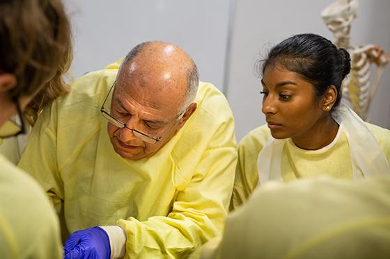 Photo of an instructor wearing yellow protective smock, leaning over cadaver while Chatham University students watch on. 